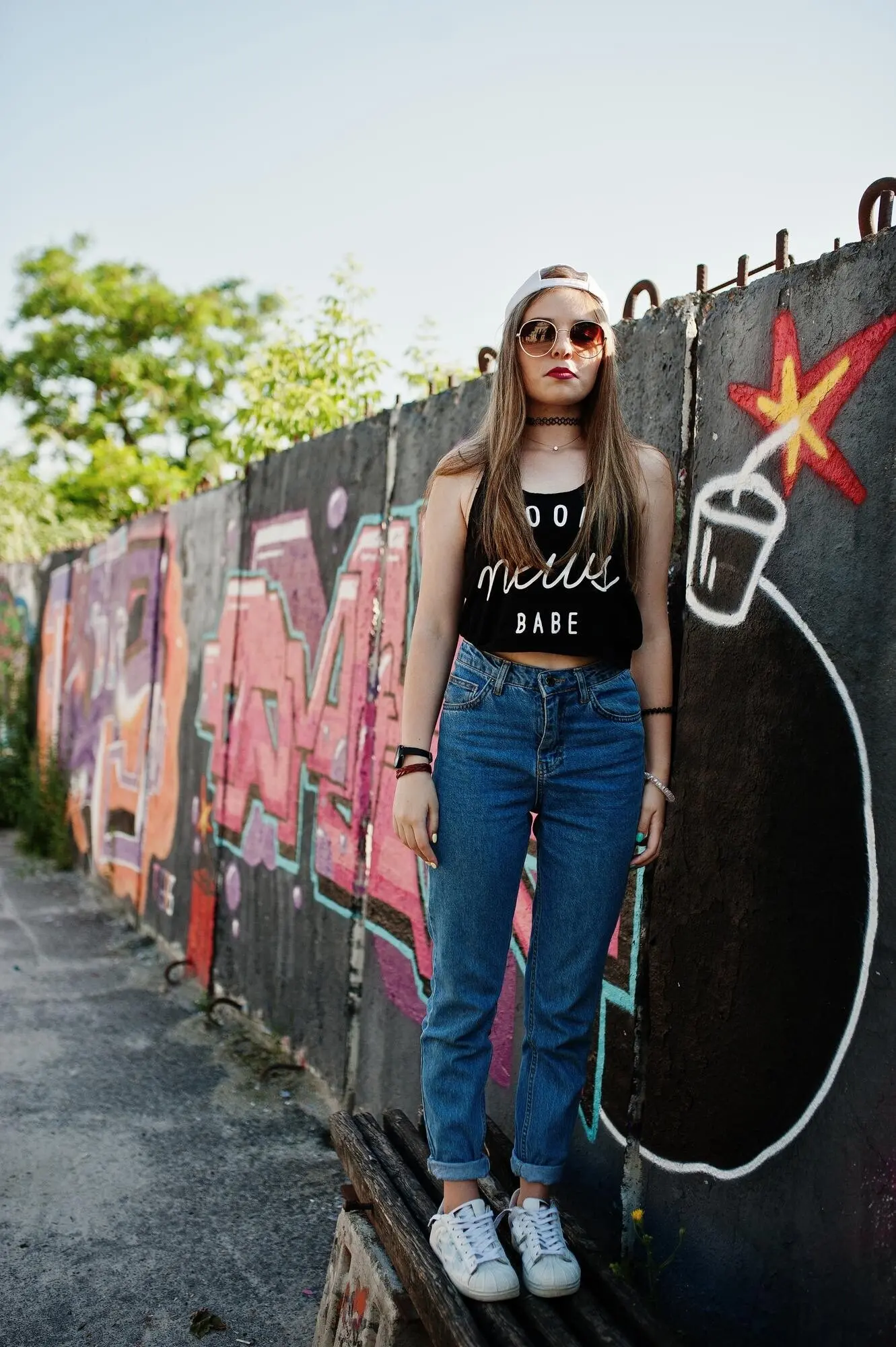 Chica hipster casual y a la moda con gorra, gafas de sol y vaqueros, frente a una gran pared de grafiti con una gran bomba de TNT.